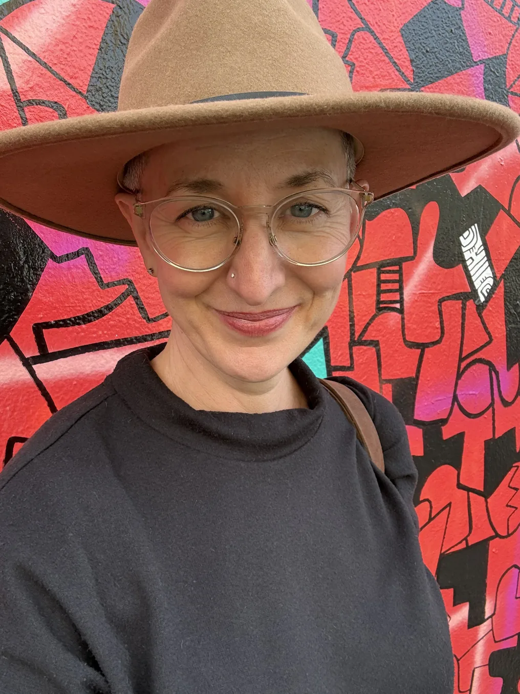 Portrait of Chrissie Ott smiling, wearing a wide-brimmed hat, standing in front of a colorful mural