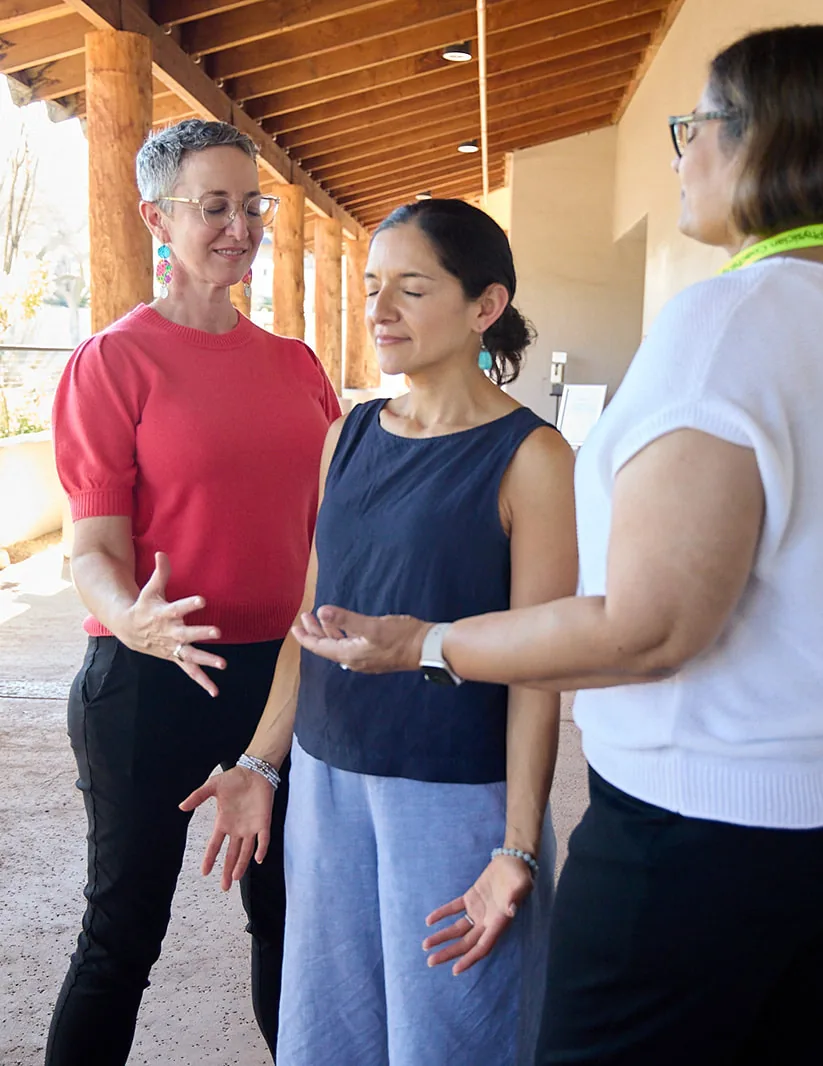 Three women in a moment of guided touch and presence, eyes closed, hands extended